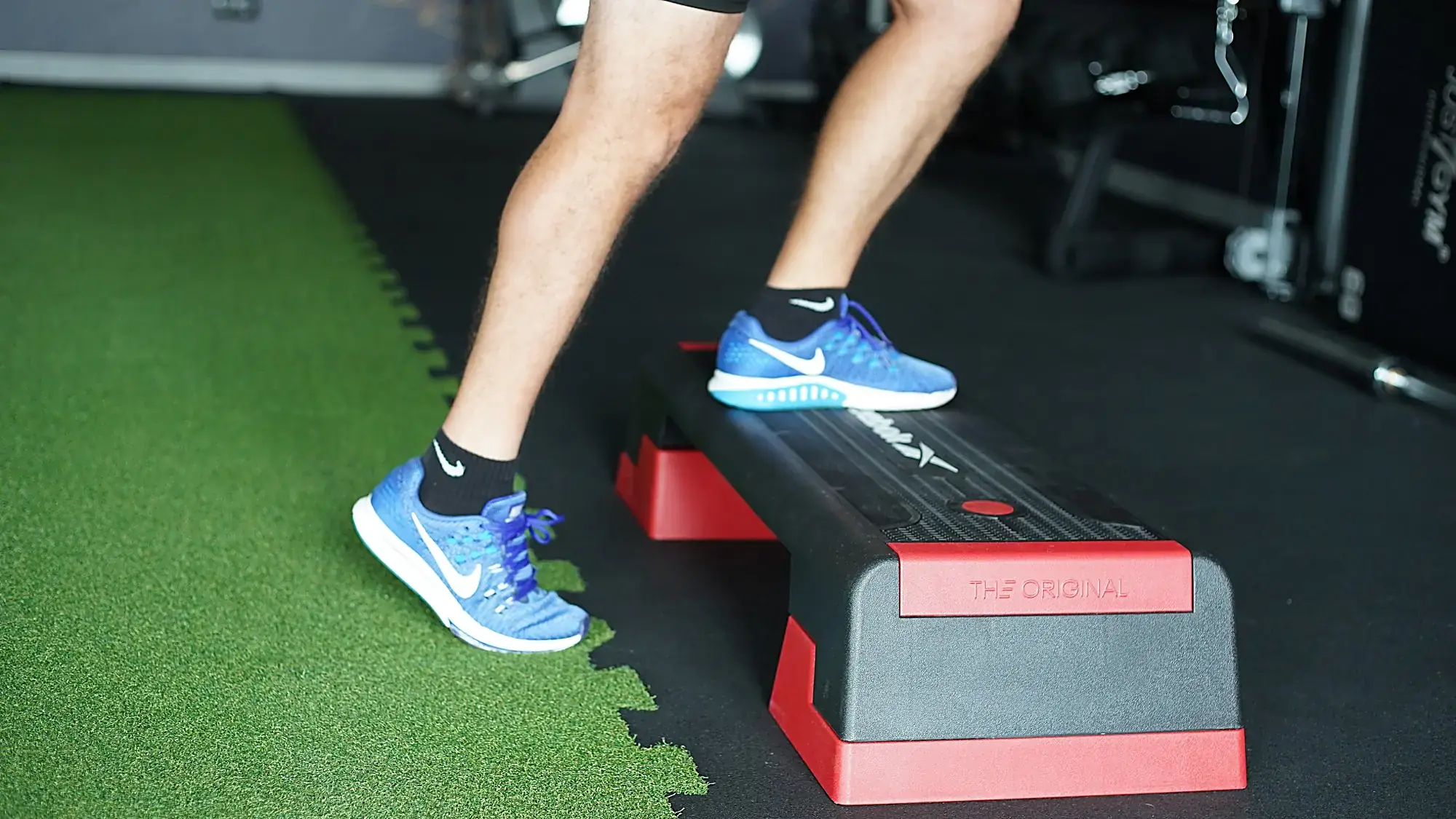 A man in blue running shoes doing a calf raise on an aerobic step in the Teho Sport training room, with gym machines behind him and a strip of artificial turf beside